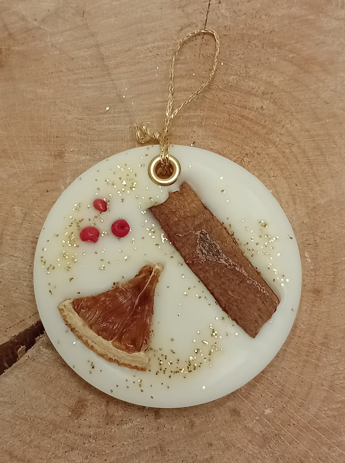 Circular wax sachet decorated with a cinnamon stick, orange slice, pink peppercorns and gold glitter with a wooden log slice background.