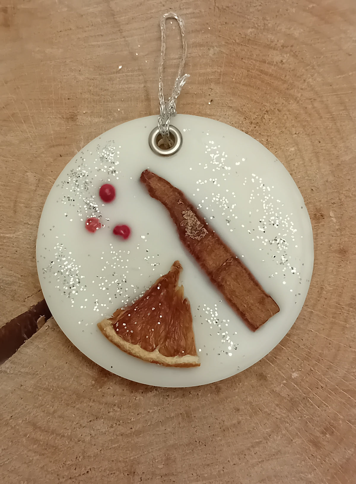Circular wax sachet decorated with a  cinnamon stick, orange slice, pink peppercorns and silver glitter with a wooden log slice background.