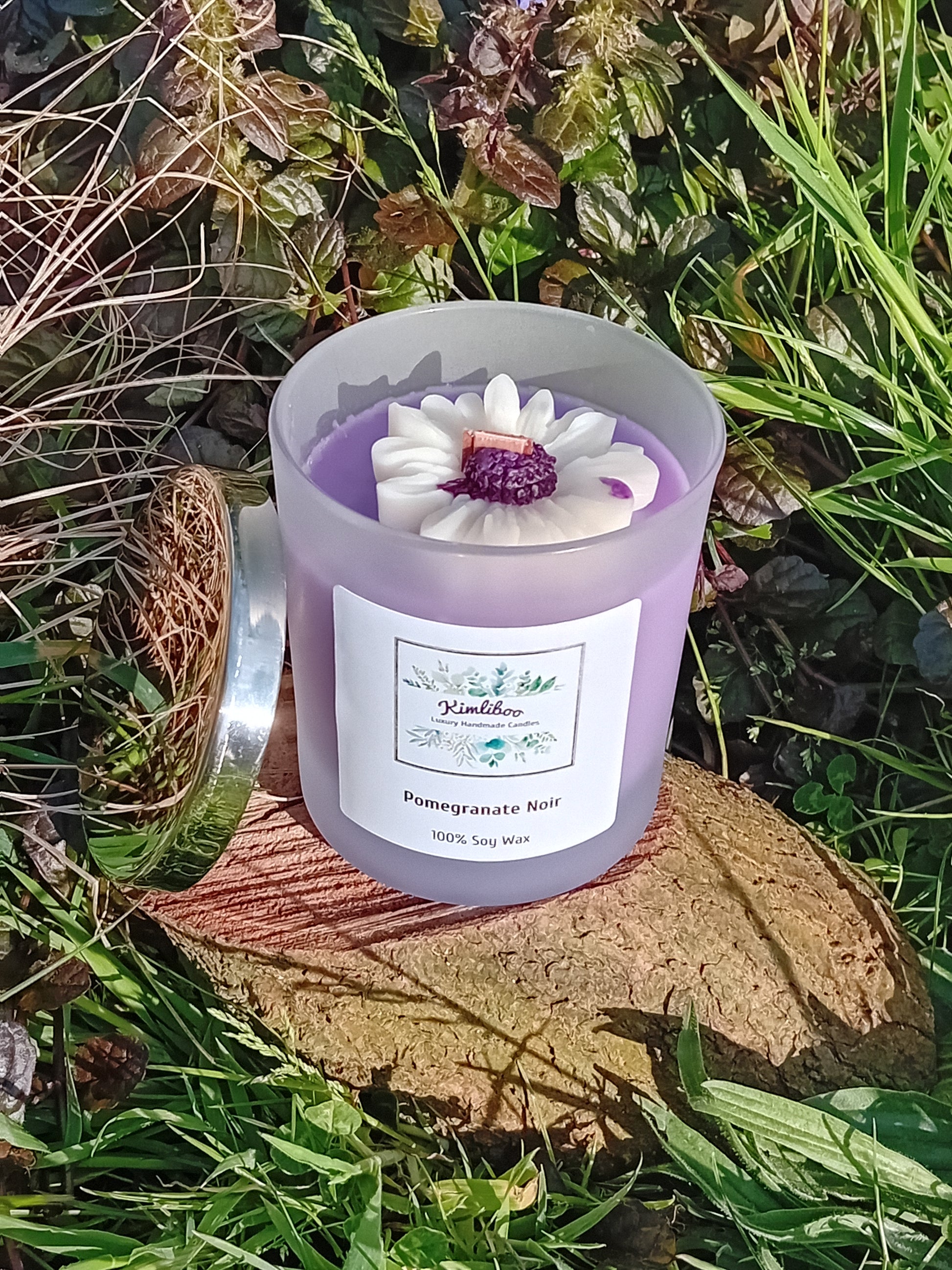 Pomegranate Noir fragranced purple candle in a 30cl frosted jar with wooden wick and a large daisy-shaped wax decoration. 