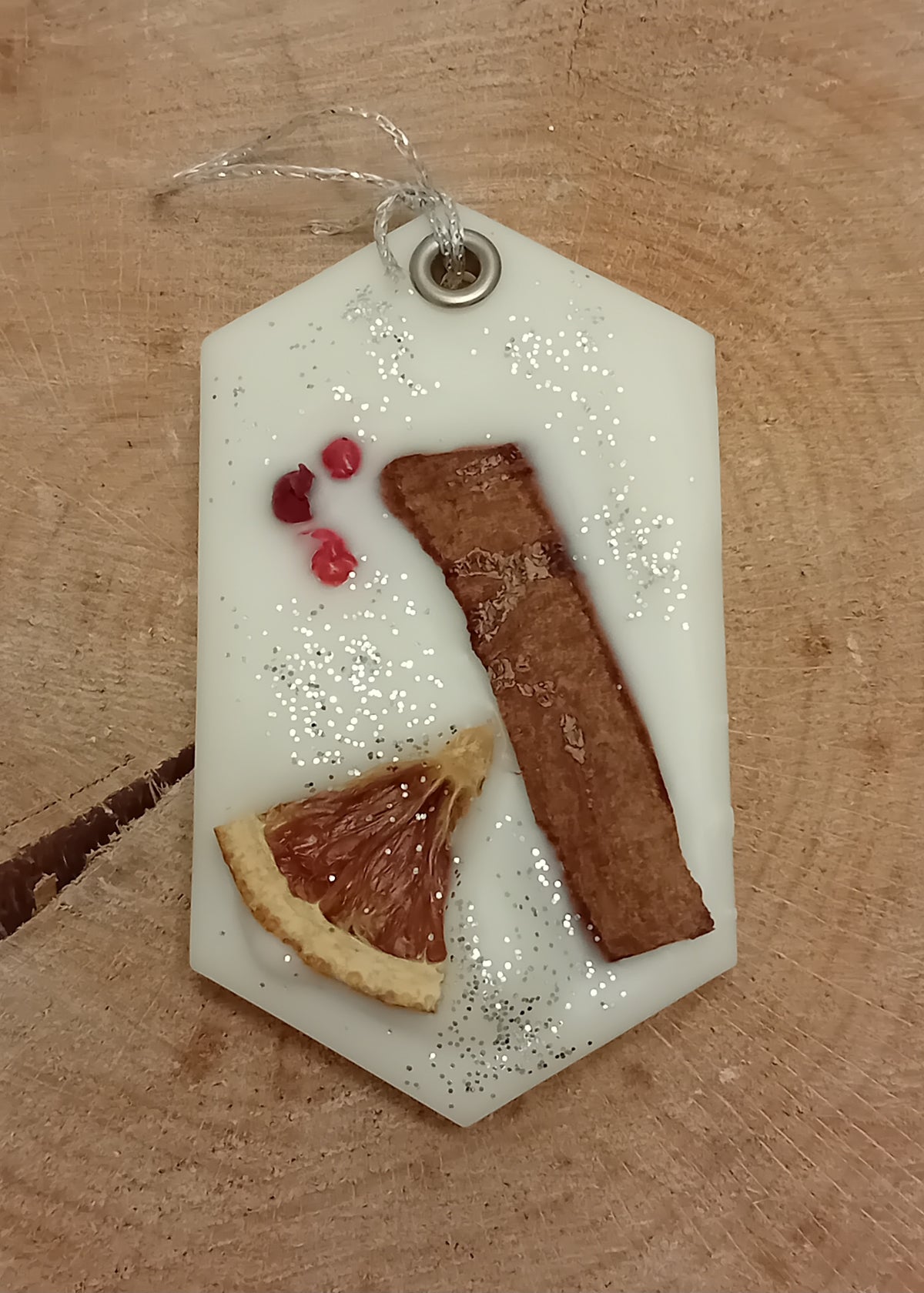 Hexagonal wax sachet decorated with a cinnamon stick, orange slice, pink peppercorns and silver glitter with a wooden log slice background.