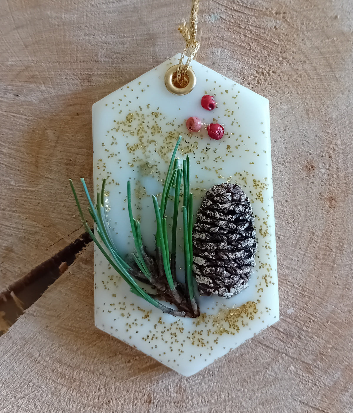 Hexagonal wax sachet decorated with a birch pine cone, pine tree leaves, pink peppercorns and gold glitter with a wooden log slice background.