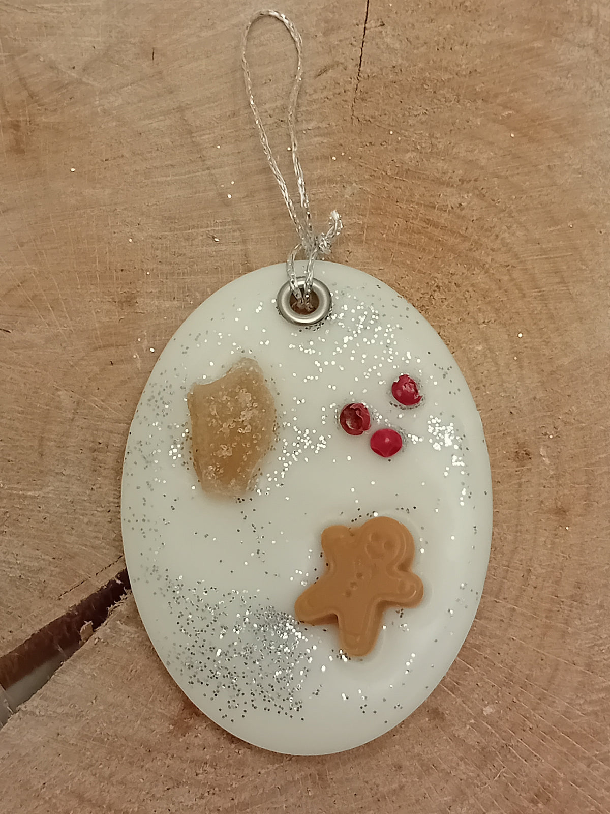 Oval wax sachet decorated with a piece of crystallized ginger, a wax gingerbread man, pink peppercorns and silver glitter with a wooden log slice background.