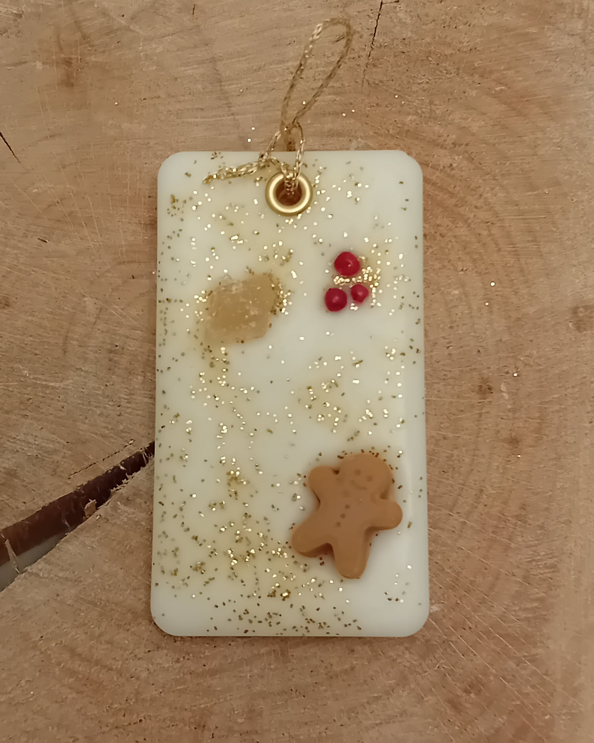 Rectangular wax sachet decorated with a piece of crystallized ginger, a wax gingerbread man, pink peppercorns and gold glitter with a wooden log slice background.