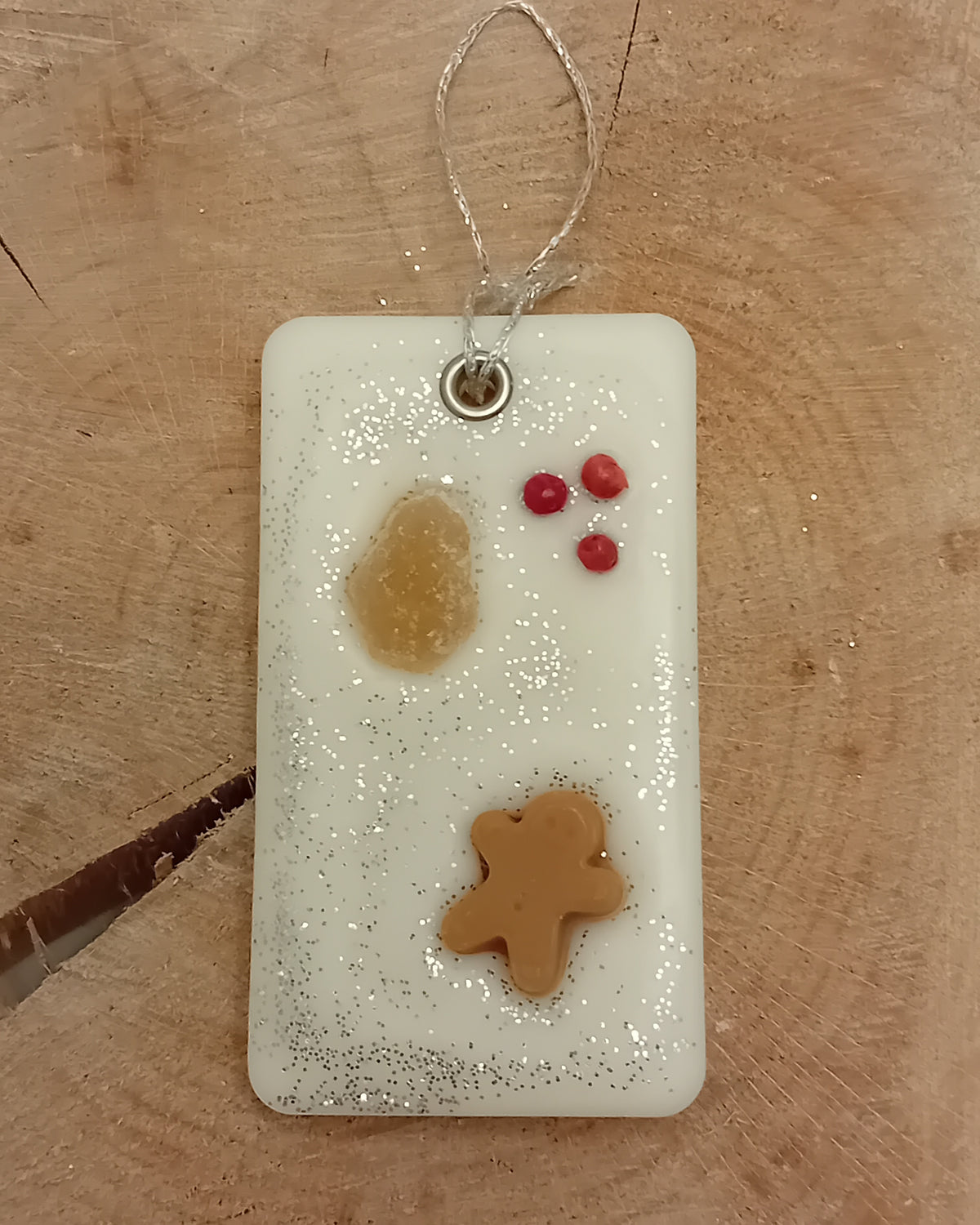 Rectangular wax sachet decorated with a piece of crystallized ginger, a wax gingerbread man, pink peppercorns and silver glitter with a wooden log slice background.