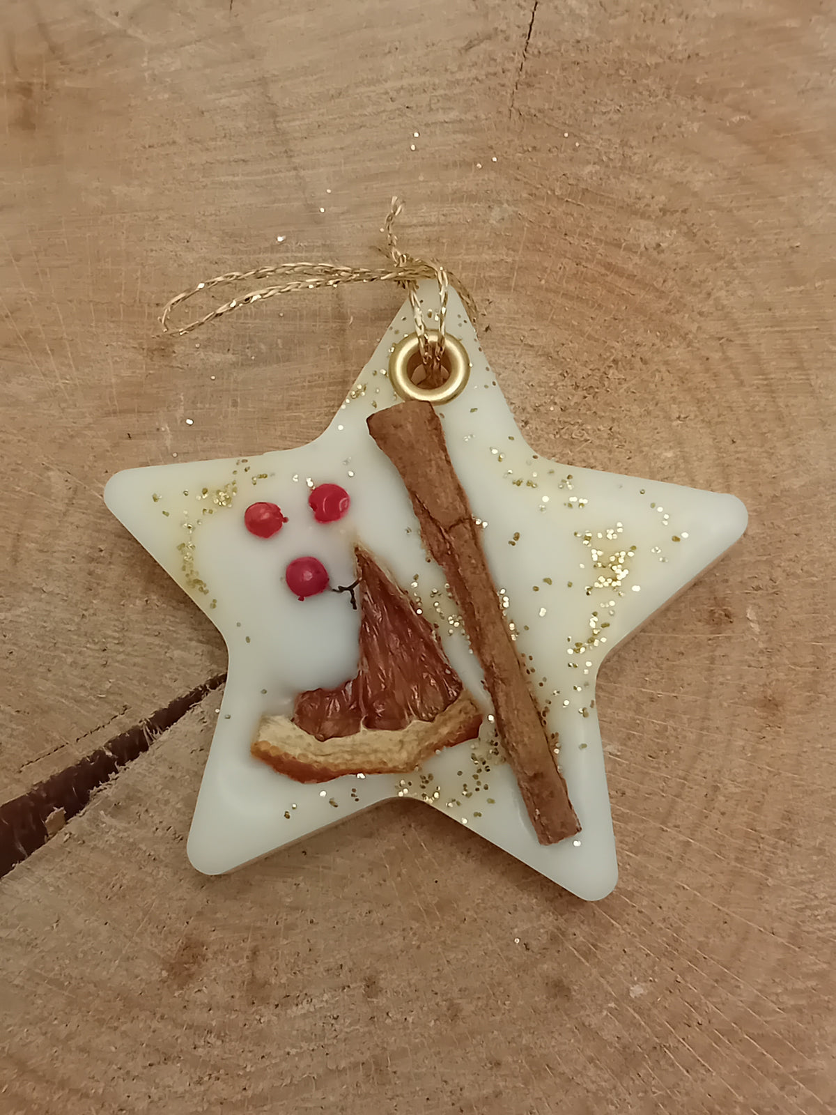 Star-shaped wax sachet decorated with a cinnamon stick, orange slice, pink peppercorns and gold glitter with a wooden log slice background.