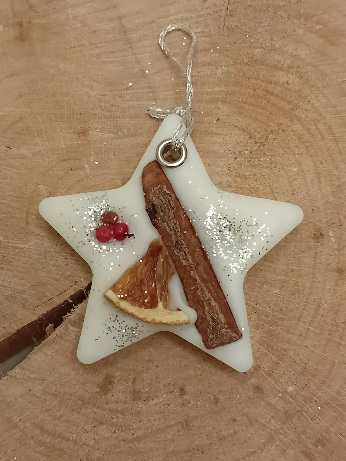 Star-shaped wax sachet decorated with a cinnamon stick, orange slice, pink peppercorns and silver glitter with a wooden log slice background.