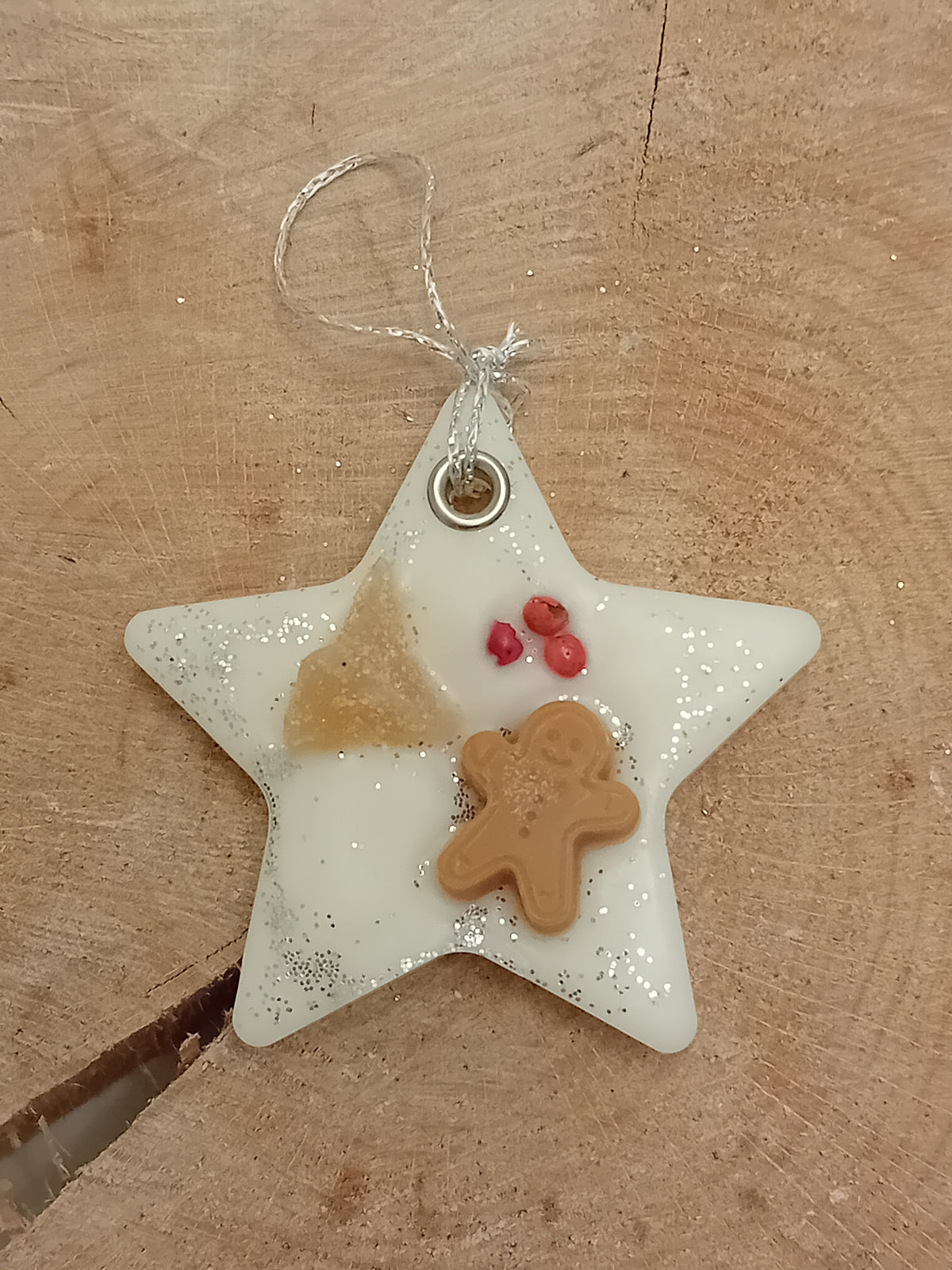 Star-shaped wax sachet decorated with a piece of crystallized ginger, a wax gingerbread man, pink peppercorns and silver glitter with a wooden log slice background.